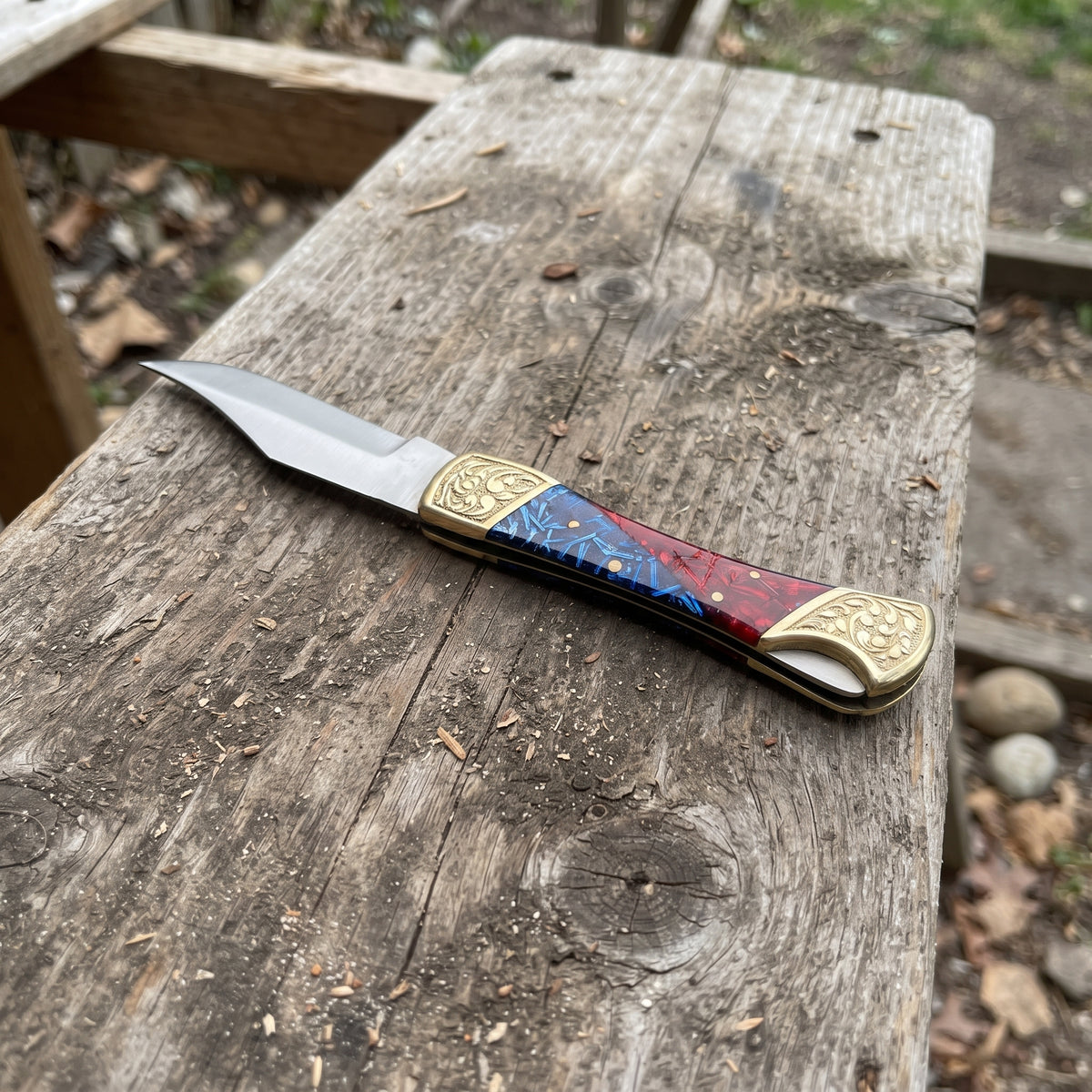 A pocket knife with a steel blade and a decorative handle featuring red, blue, and gold inlay, resting on a weathered wooden plank outdoors.