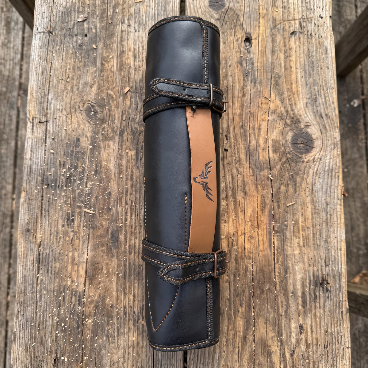 A rolled black leather knife sheath with a tan leather accent stripe and a deer head logo, displayed on a rustic wooden plank background.