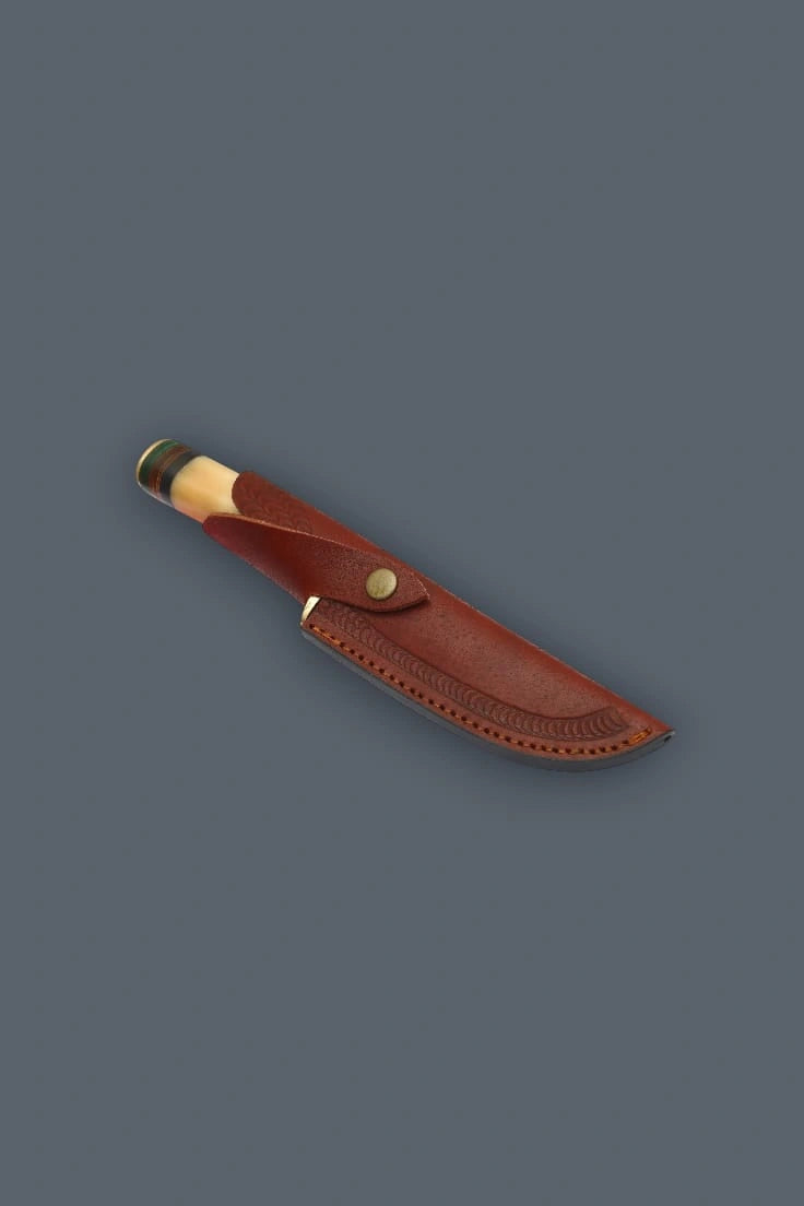 Diagonal flat lay of the Damascen Knives fixed blade hunting knife secured in its genuine leather sheath, highlighting the stamped edge tooling and brass snap.