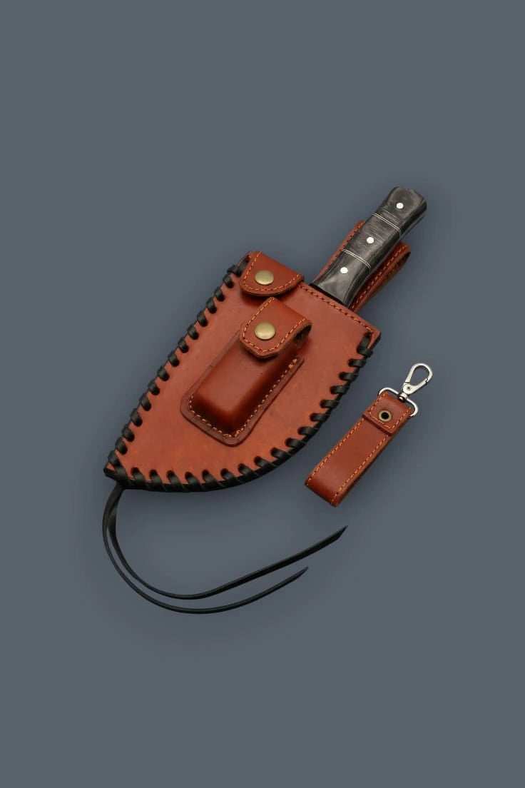 Diagonal flat lay of the Damascus meat cleaver secured in its heavily laced leather sheath, featuring a front utility pocket, brass snaps, and a detached leather clip.