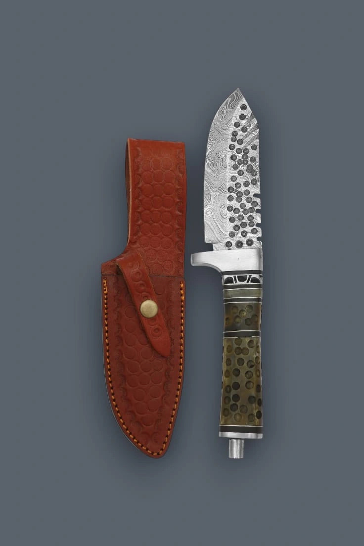 Diagonal flat lay of the Damascen Knives hunting knife secured in its genuine leather sheath, highlighting the stamped circular pattern and brass snap closure.