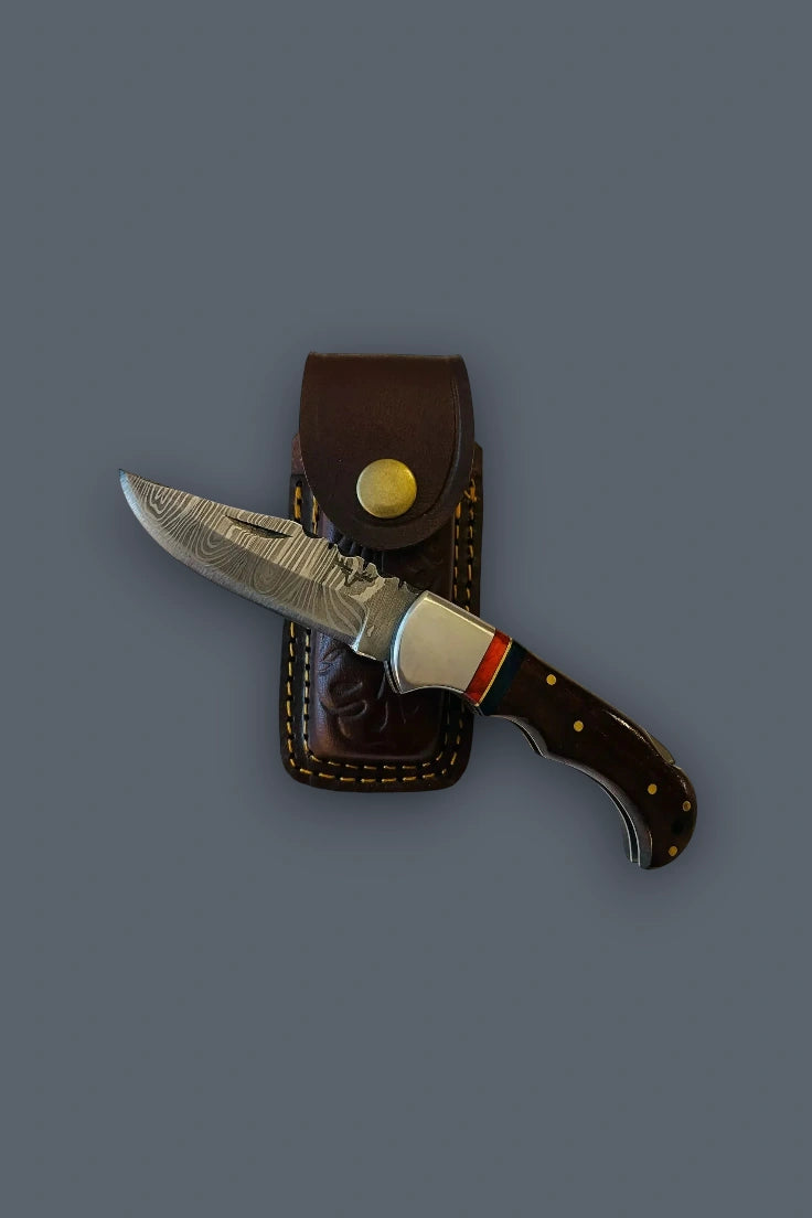 Flat lay of the fully open The Gentry Knife resting horizontally across its closed brown tooled leather sheath, emphasizing the Damascus blade profile.