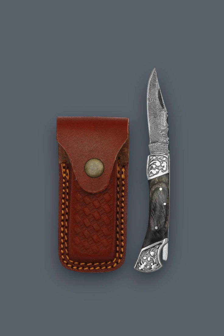 Vertical flat lay of the fully open The Squire Damascus compact folding pocket knife with sheath positioned alongside its closed brown basketweave leather sheath