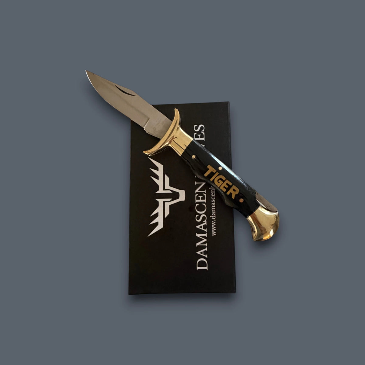 A Damascus steel fixed-blade knife with a black handle and gold accents, resting diagonally on top of its black branded presentation card against a solid dark gray background.