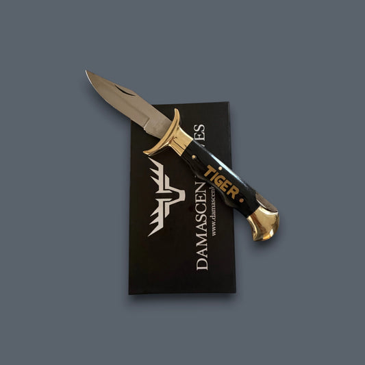 A Damascus steel fixed-blade knife with a black handle and gold accents, resting diagonally on top of its black branded presentation card against a solid dark gray background.