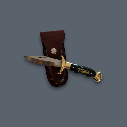 A fixed-blade knife with a black handle, gold guard and pommel, and the word 'TIGER' printed on the handle, shown next to a brown leather sheath on a solid dark gray background.
