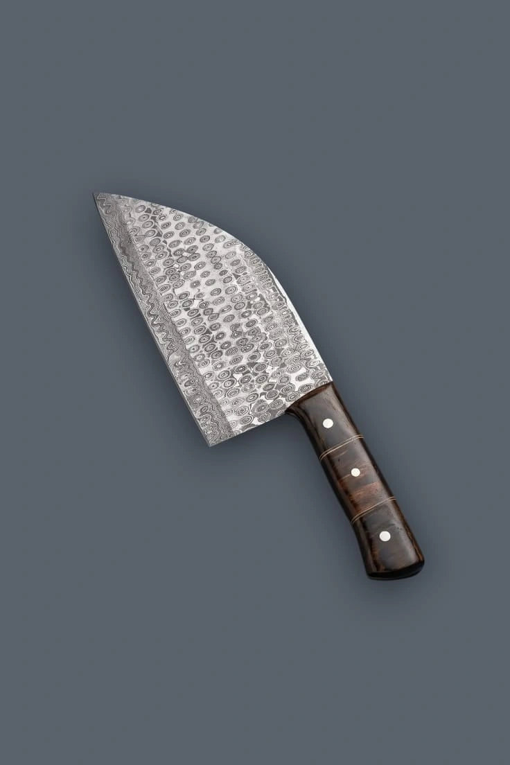 Upward diagonal flat lay of the Damascus meat cleaver, highlighting the unique raindrop steel pattern and the contoured dark Pakkawood grip with metal spacers.